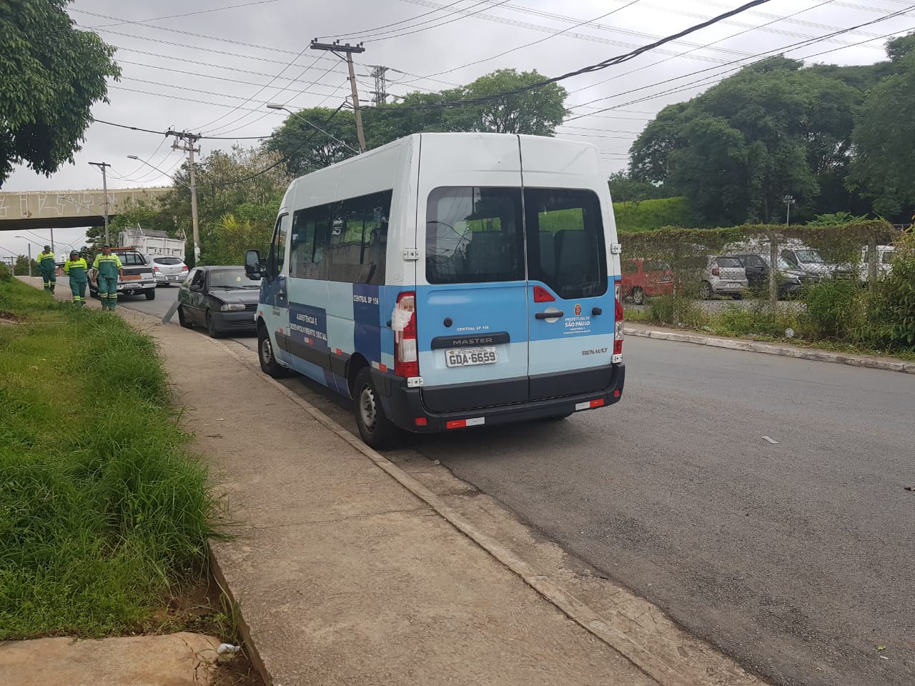 Via urbana próxima ao incêndio com uma van estacionada, carros, homens uniformizados em movimento na calçada, veículo da defesa civil ao fundo 